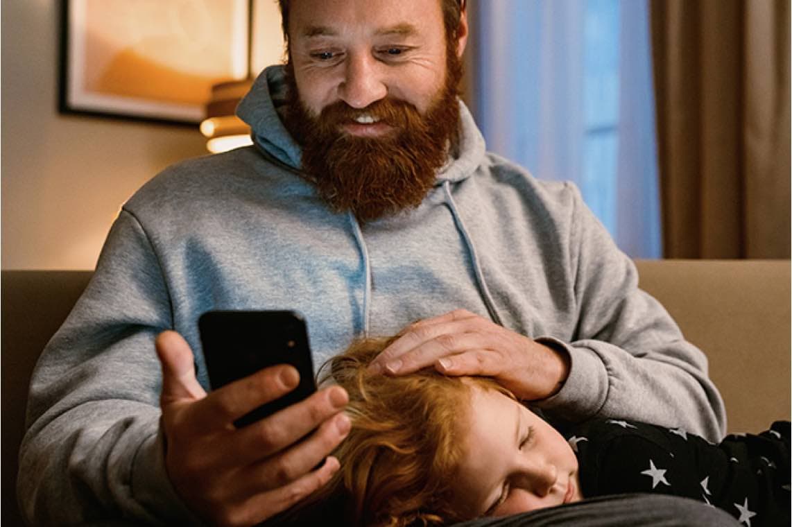 Mann mit Bart sitzt auf dem Sofa, schaut auf sein Smartphone und streicht einem Kind über den Kopf