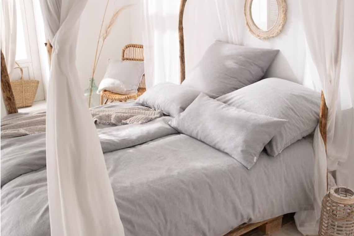 Doppelbett mit grauer Bettwäsche und vielen Kissen, umgeben von weißen Vorhängen in hellem Schlafzimmer