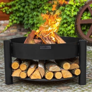 Runde Feuerstelle mit brennendem Holz, darunter ordentlich gestapelte Holzscheite im Garten vor grüner Hauswand