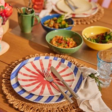 Gedeckter Tisch mit bunten Tellern, Besteck, Trinkglas und Schalen mit Speisen auf geflochtenen Platzsets