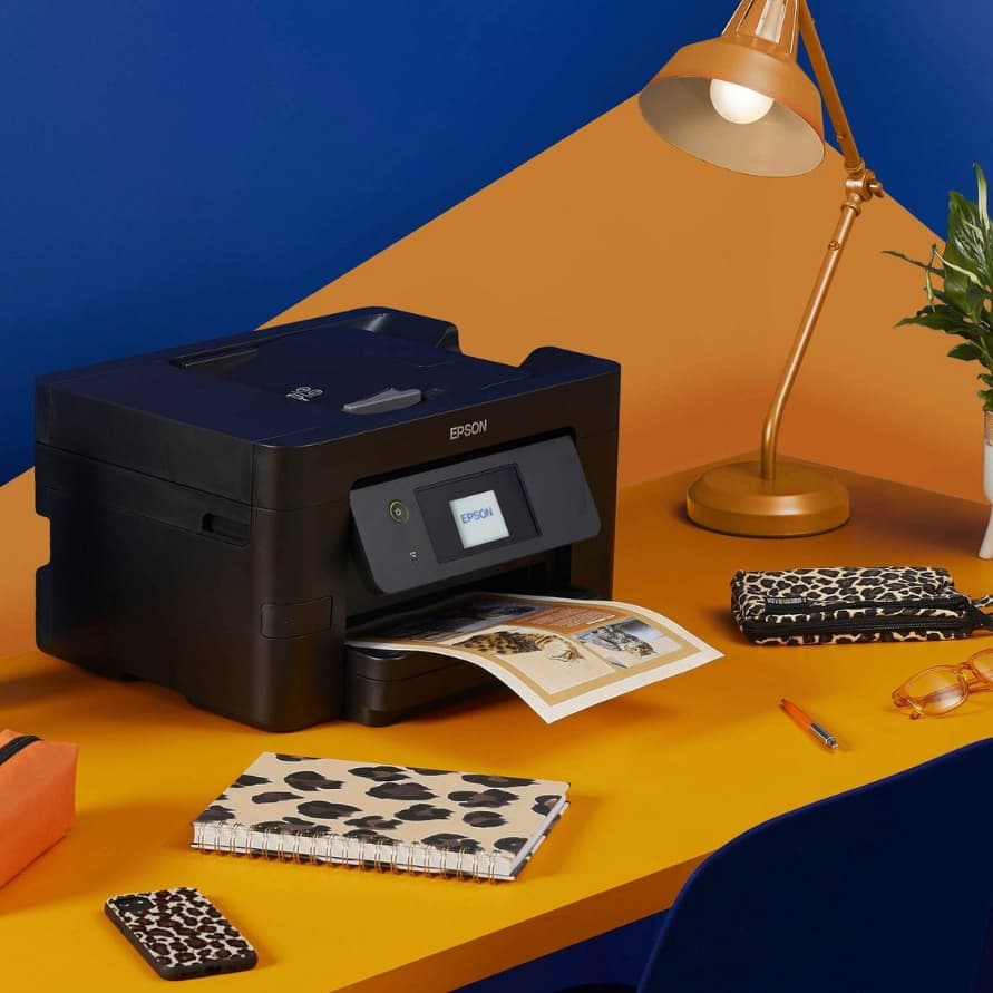 Schwarzer Epson-Drucker auf Schreibtisch, druckt Foto, daneben Lampe, Notizbuch, Handyhülle und Stifte