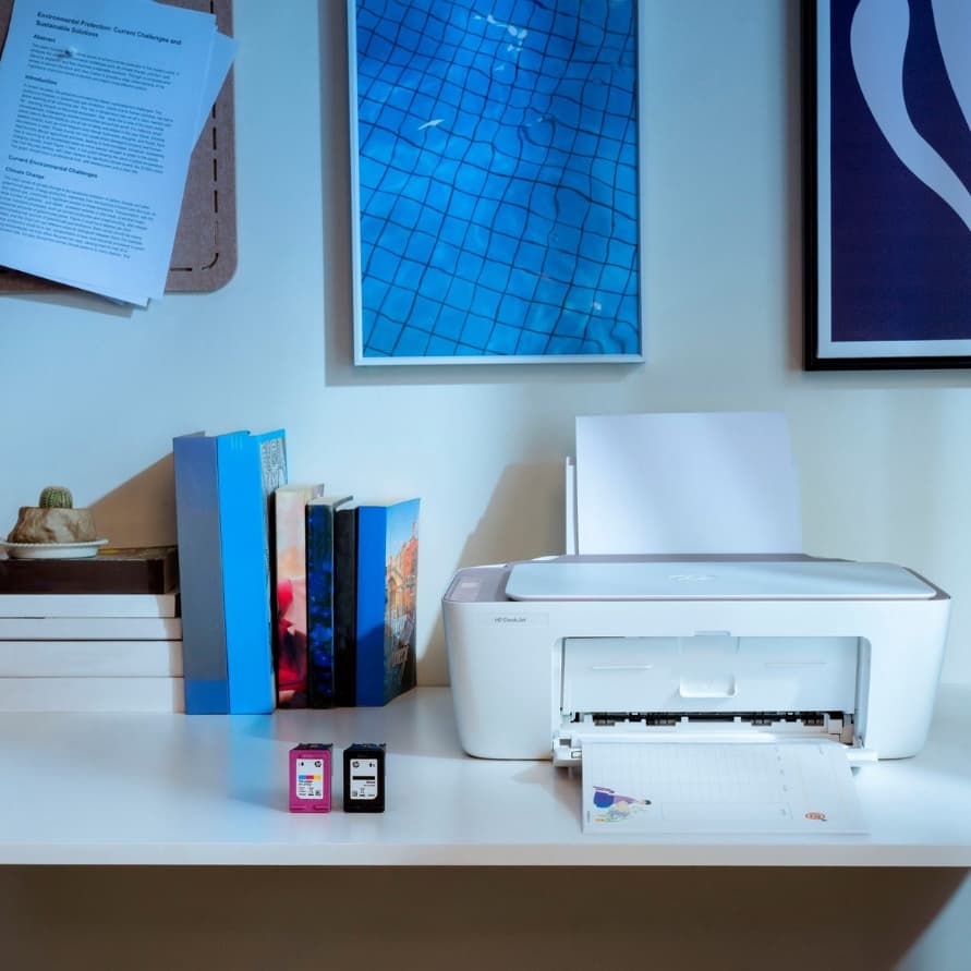 Weißer Drucker auf Schreibtisch druckt ein Blatt, daneben zwei Patronen, Bücher und Bilder an der Wand