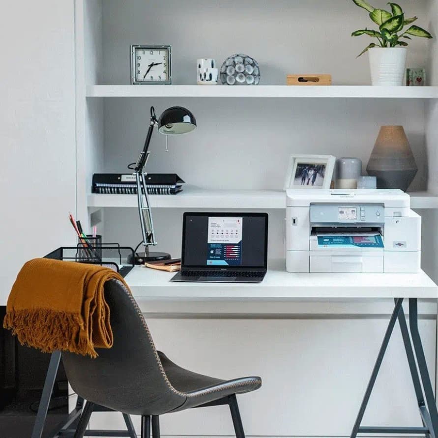 Schreibtisch mit Laptop, Drucker, Schreibtischlampe und Stuhl mit Tuch, dahinter weiße Regale mit Uhr, Ordnern und Pflanze