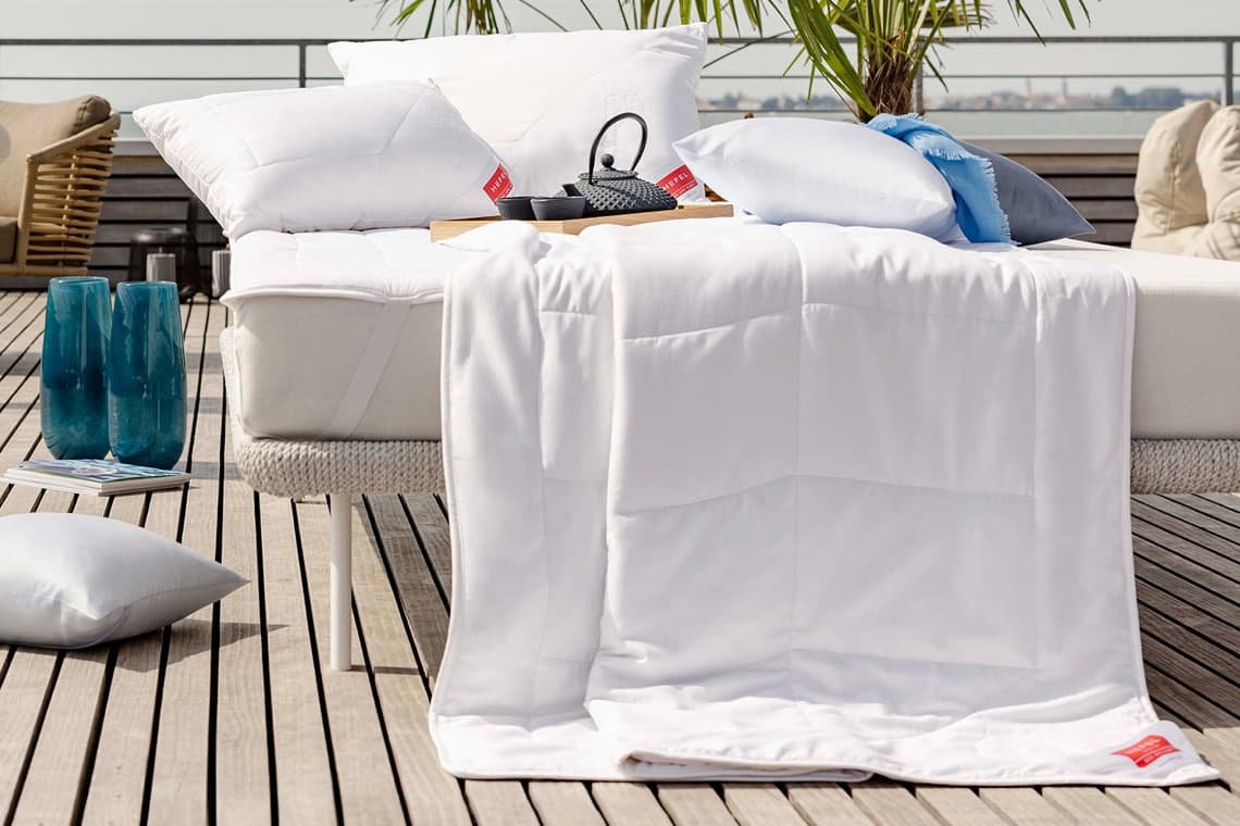 Weiße Bettdecke mit passenden Kissen auf Terrasse, daneben blaue Vasen, Kissen auf Holzboden und Palmen im Hintergrund