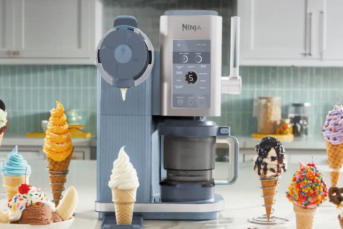 Eismaschine auf Küchenarbeitsplatte, vorne verschiedene bunte Eistüten mit Softeis und Toppings