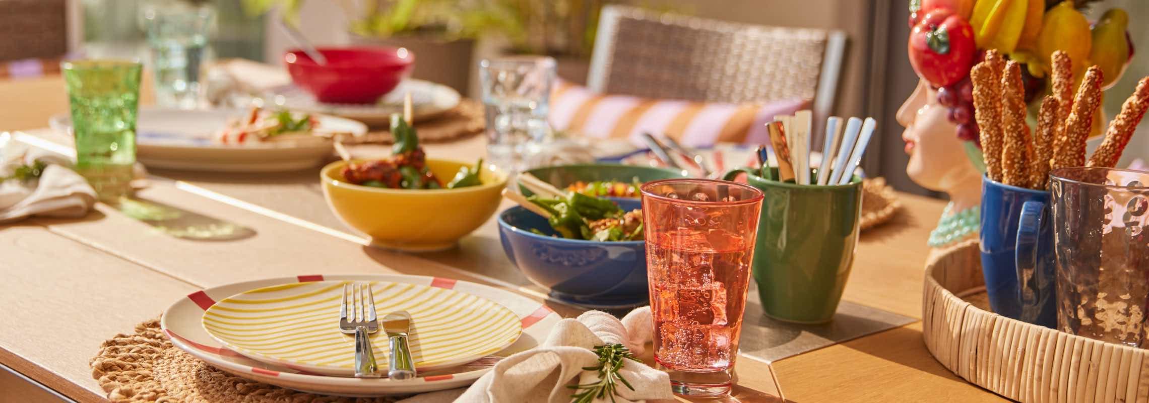 Bunt gedeckter Gartentisch mit Tellern, Gläsern, Schalen mit Speisen und Brotstangen in Bechern