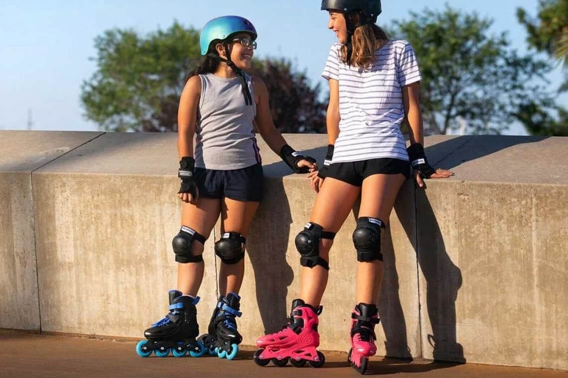 Zwei Mädchen mit Helm und Schutzausrüstung stehen auf Inlineskates an einer Mauer und halten sich an den Händen