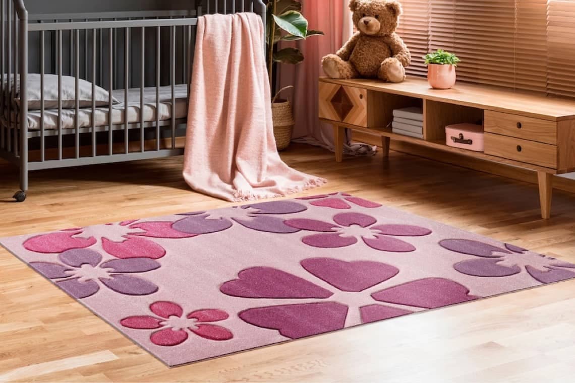 Rosa Kinderzimmerteppich mit großen Blumen- und Herzmotiven vor Babybett und Lowboard mit Teddybär