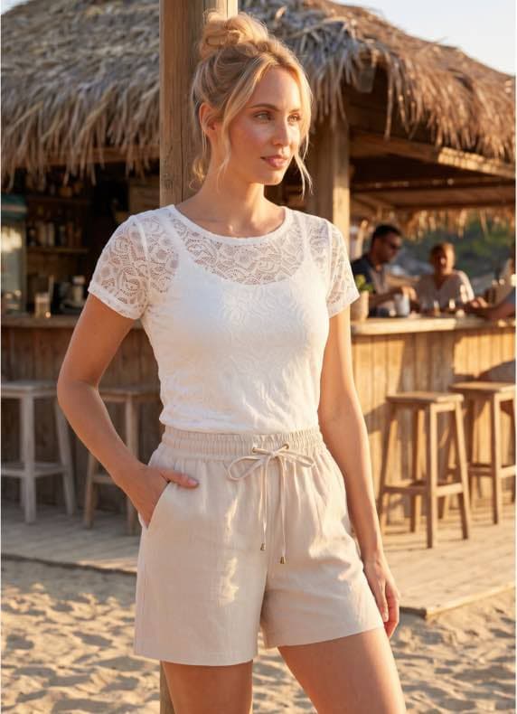 Frau mit weißem Spitzenshirt und hellen Shorts steht am Strand vor einer Strandbar und schaut zur Seite