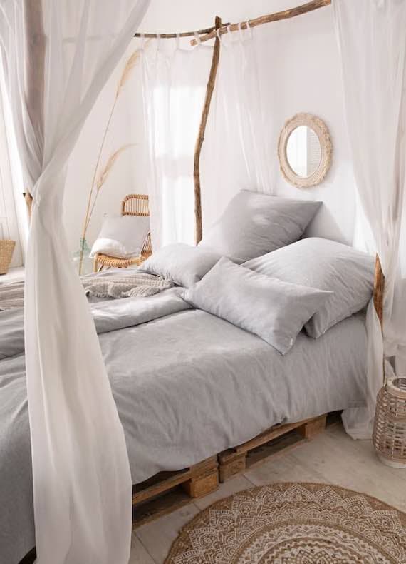 Himmelbett mit grauer Bettwäsche auf Paletten, umgeben von weißen Vorhängen, Korbstuhl und rundem Teppich im Schlafzimmer
