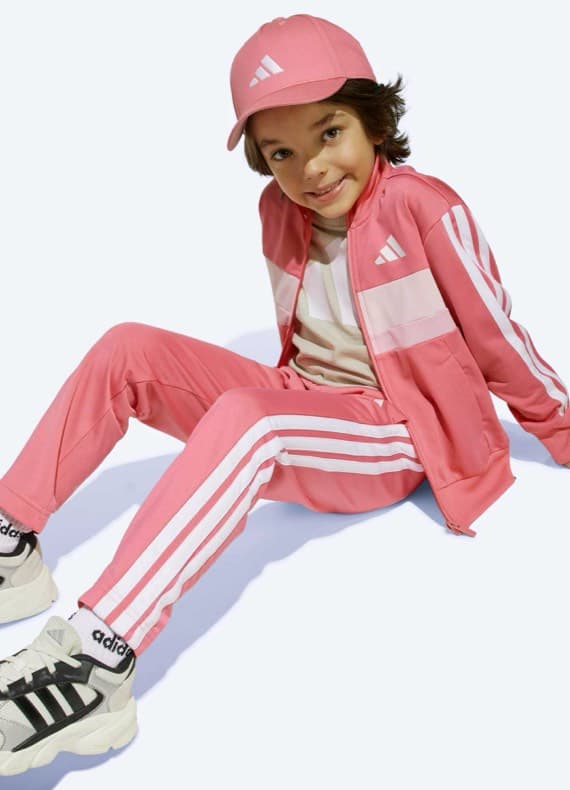 Kind sitzt entspannt auf dem Boden und trägt pinken Trainingsanzug, Kappe und Turnschuhe mit drei Streifen