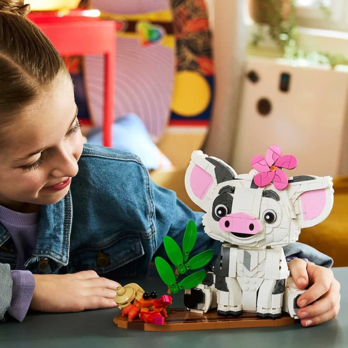 Mädchen stützt sich auf den Tisch und spielt mit einem bunten Lego-Ferkel mit Blume im Haar und kleiner Dschungelszene