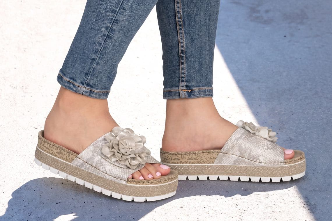 Füße in hellen Plateausandalen mit großer Blüte, getragen zu hochgekrempelter blauer Jeans auf hellem Boden