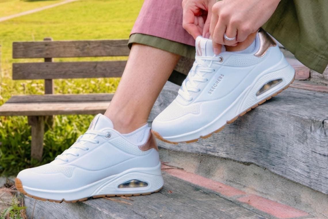 Person sitzt auf Bank im Park und schnürt weiße Skechers-Sneaker mit dicker Sohle und sichtbarem Luftpolster