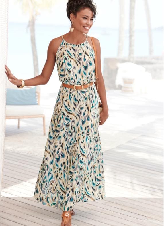 Frau im ärmellosen Maxikleid mit buntem Muster und Gürtel geht lächelnd über eine helle Strandterrasse