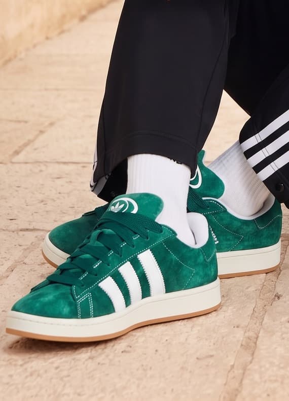Person sitzt auf Steinboden, trägt grüne Sneakers mit drei weißen Streifen, weiße Socken und schwarze Trainingshose
