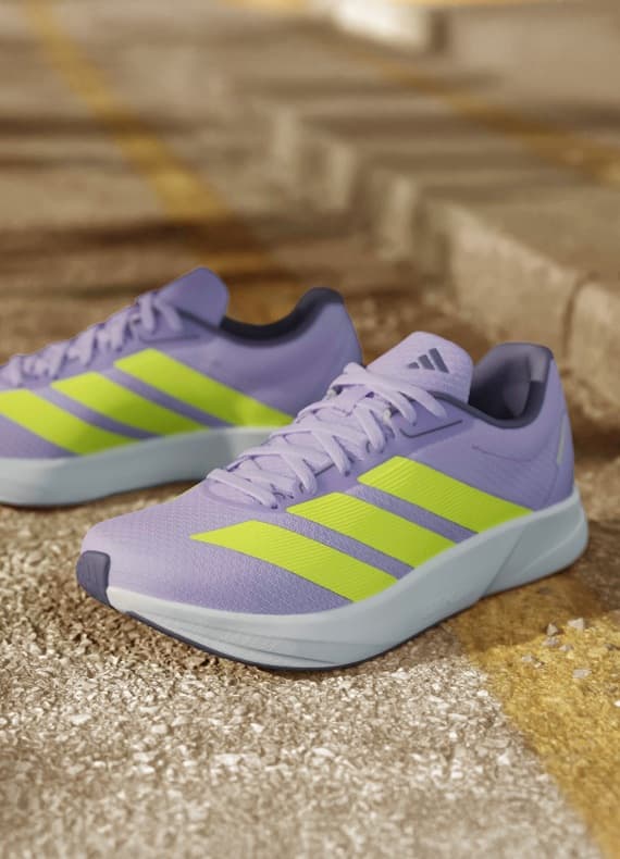 Fliederfarbene Adidas-Sneaker mit gelben Streifen stehen nebeneinander auf einem sonnigen Asphaltweg