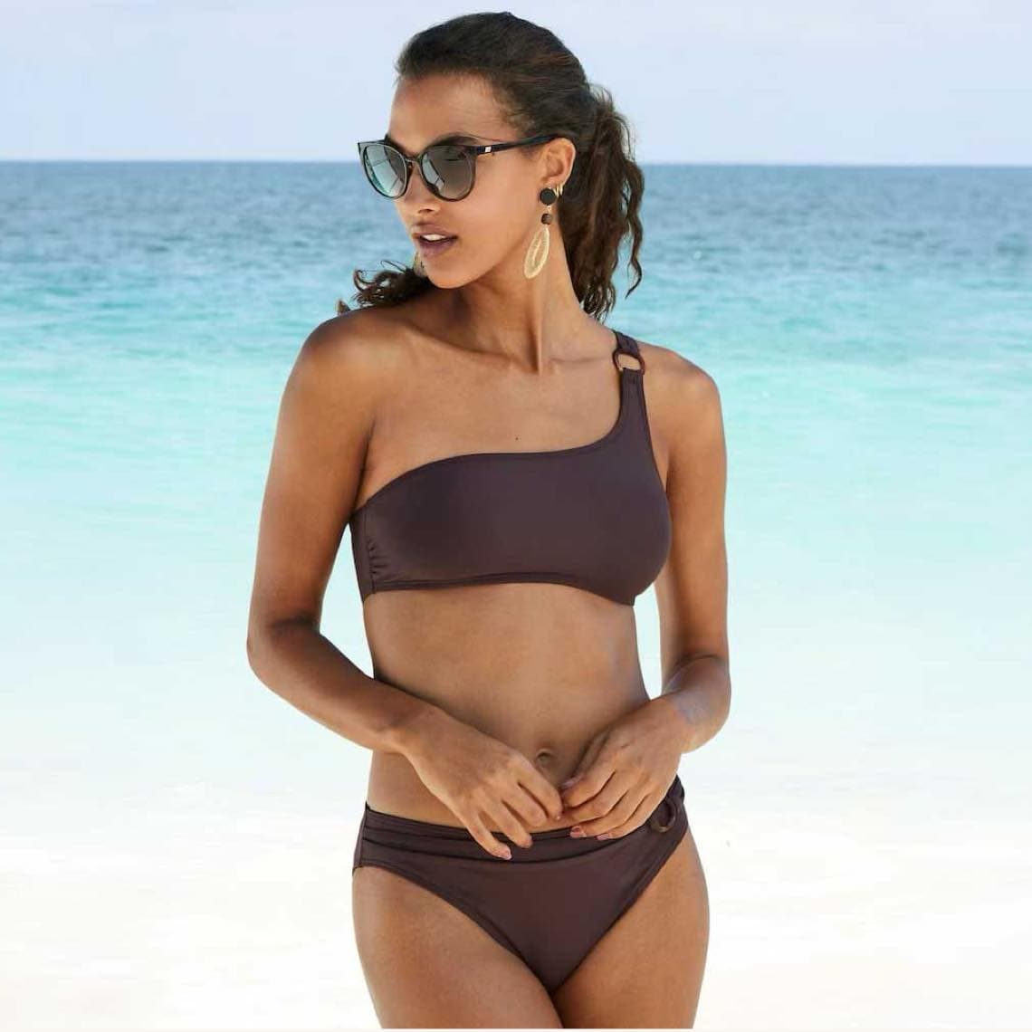 Frau im einseitigen Bikini mit Sonnenbrille steht am Strand vor türkisblauem Meer