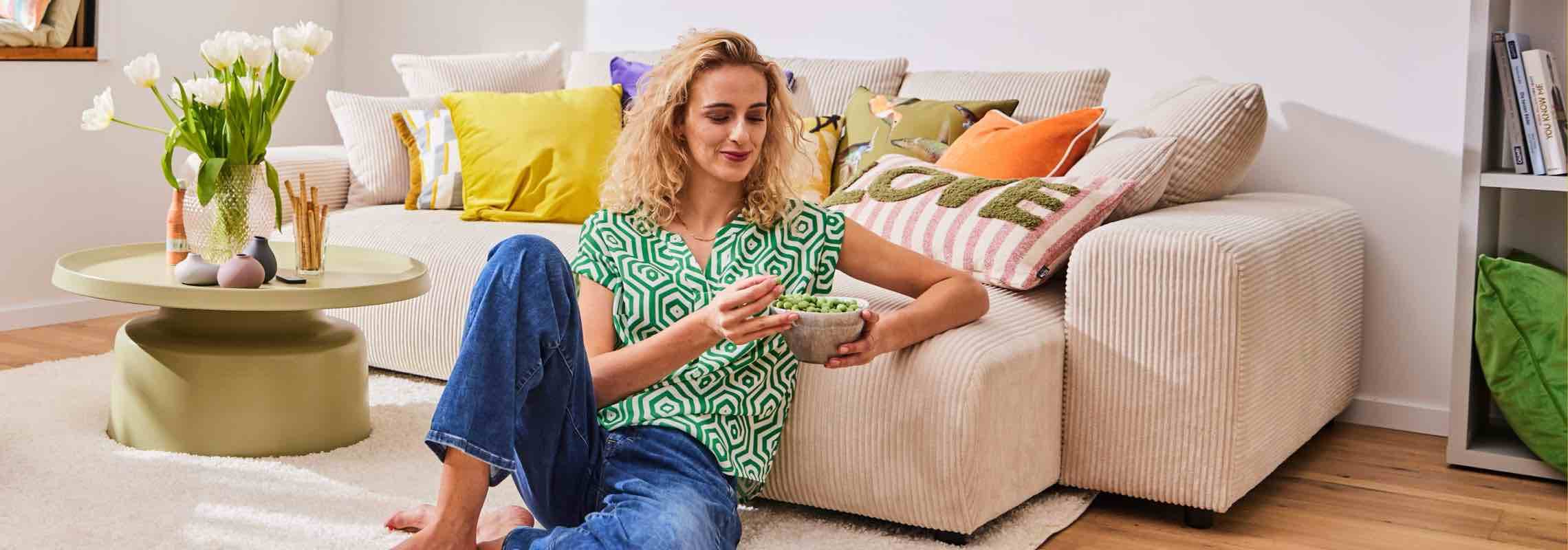 Frau sitzt barfuß vor beigem Sofa und isst aus einer Schale, bunte Kissen und Tulpen auf rundem Couchtisch