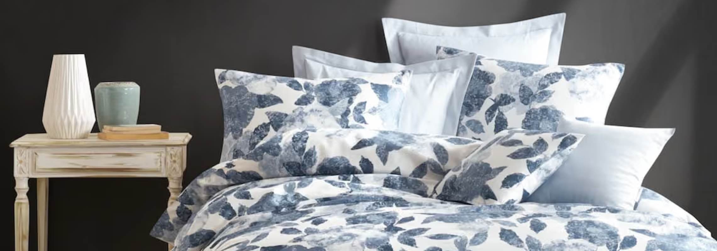 Bett mit weiß-blauer Bettwäsche im Blätterdruck, mehrere Kissen vor dunkler Wand, daneben Tisch mit Vasen