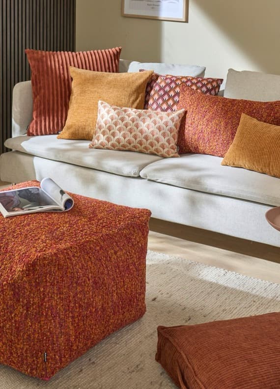 Helles Sofa mit vielen orange- und rostfarbenen Kissen, davor passender Pouf mit aufgeklapptem Magazin
