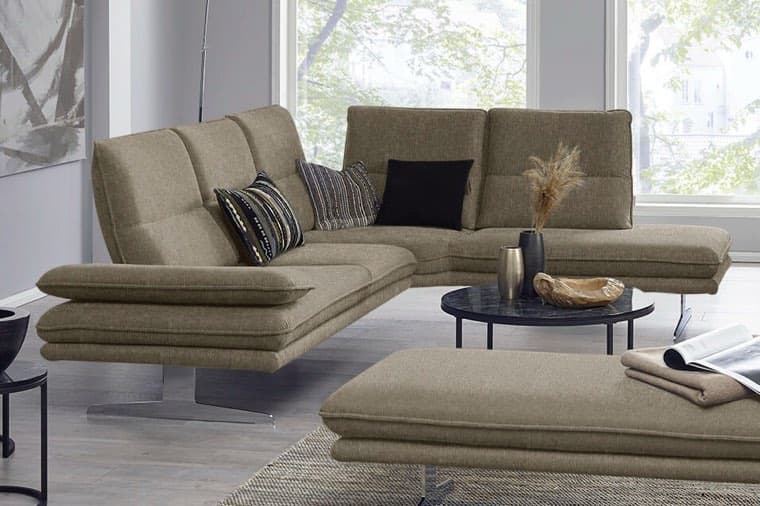 Ecksofa in Beige mit Zierkissen vor großen Fenstern, davor runder Couchtisch mit Dekovase und passender Hockerbank