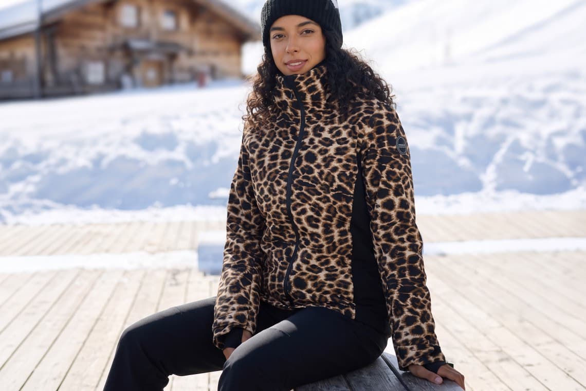 Frau in Leopardenjacke und Mütze sitzt auf Holzterrasse vor verschneiter Berghütte