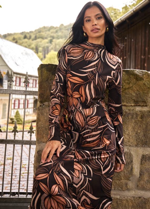 Frau in langem Kleid mit großem Blattmuster sitzt an Steinmauer vor Tor, im Hintergrund Haus und bewaldeter Hügel