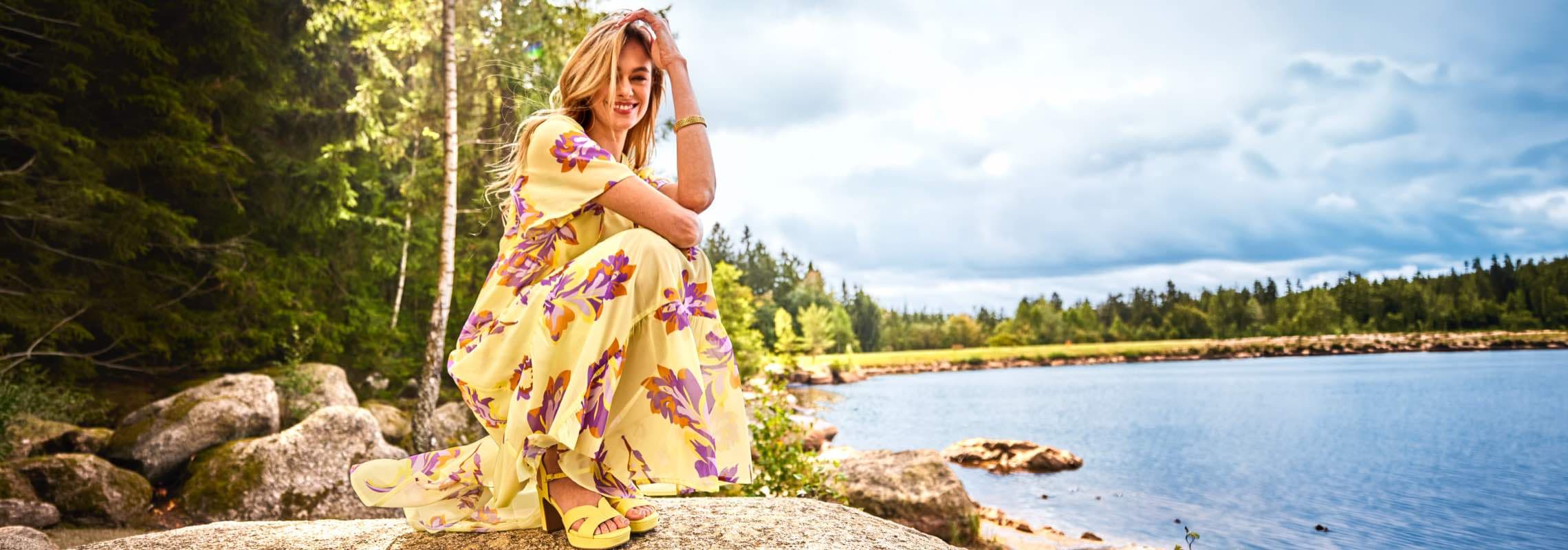 Frau im gelben, geblümten Maxikleid hockt auf Felsen am See, Wald und bewölkter Himmel im Hintergrund
