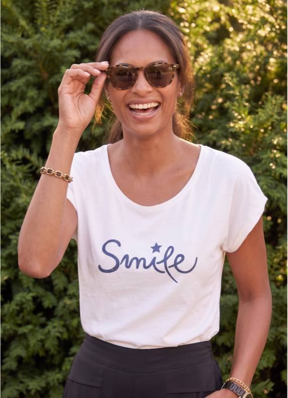Smile – Frau im weißen T-Shirt mit Schriftzug lächelt und hält Sonnenbrille, grüner Hintergrund