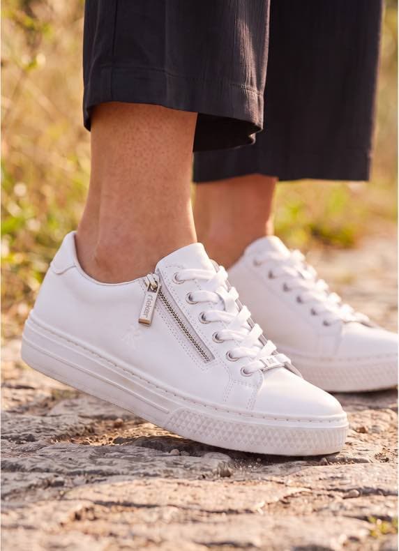 Person geht auf Steinweg, trägt weiße Sneaker mit Schnürung und seitlichem Reißverschluss unter dunkler, knöchellanger Hose
