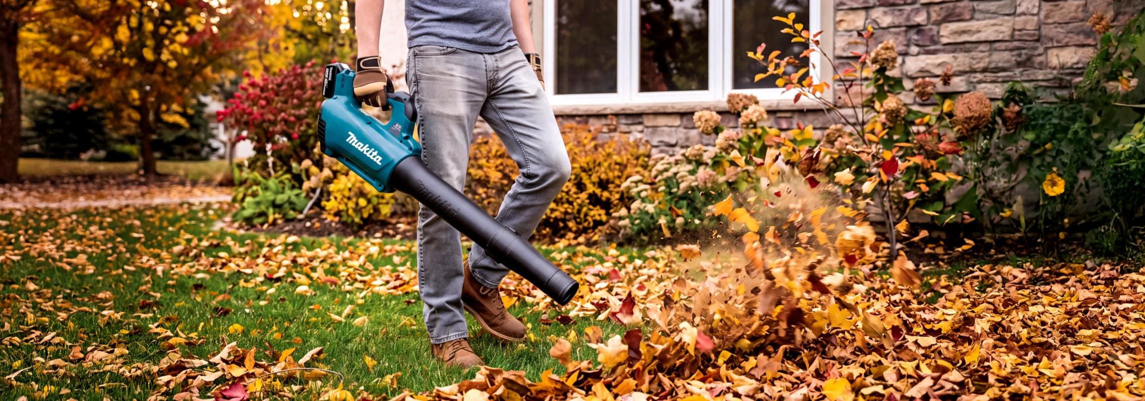 Person mit Laubbläser pustet bunte Herbstblätter von einer Rasenfläche vor einem Haus mit Garten