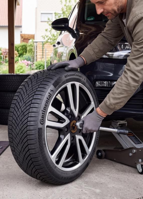 Mann wechselt Continental AllSeasonContact 2 Autoreifen an angehobenem Wagen in der Einfahrt