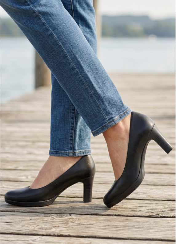 Frau in Jeans steht auf Holzsteg am Wasser und trägt schwarze Pumps mit mittelhohem Absatz