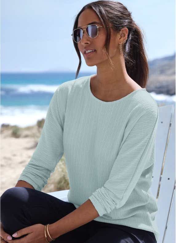Frau im mintfarbenen Langarmshirt und Sonnenbrille sitzt am Strand vor dem Meer