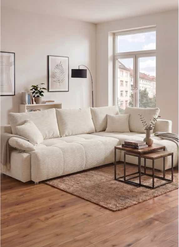 Großes helles Ecksofa im Wohnzimmer, davor Couchtisch mit Vase und Büchern auf Teppich vor Fenster mit Stadtblick