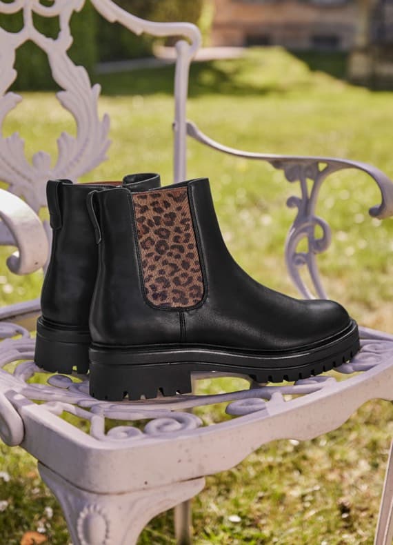 Schwarze Chelsea-Boots mit seitlichem Leopardenmuster stehen auf einem weißen Metallstuhl im Garten