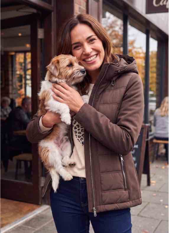 Frau in brauner Jacke steht vor Café und hält kleinen Hund im Arm