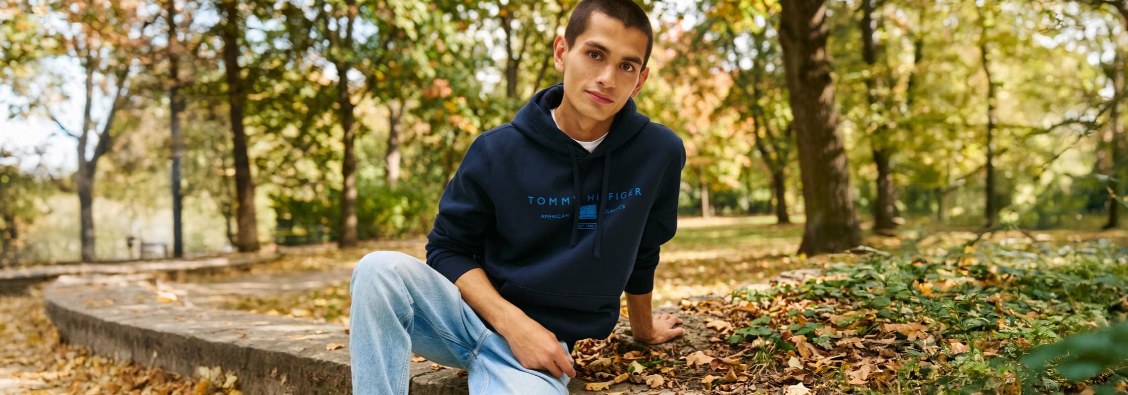 Person im dunkelblauen Hoodie mit Aufdruck sitzt im herbstlichen Park auf einer Steinmauer und schaut in die Kamera