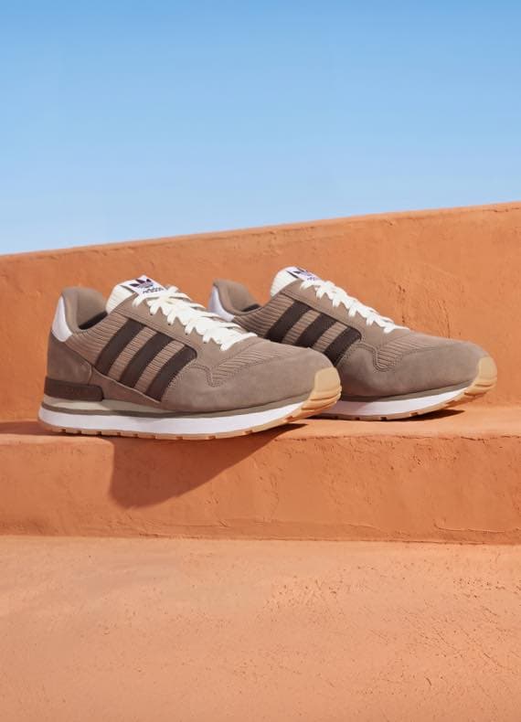 Beige Sneaker mit dunklen Streifen stehen paarweise auf terrakottafarbenen Stufen vor blauem Himmel