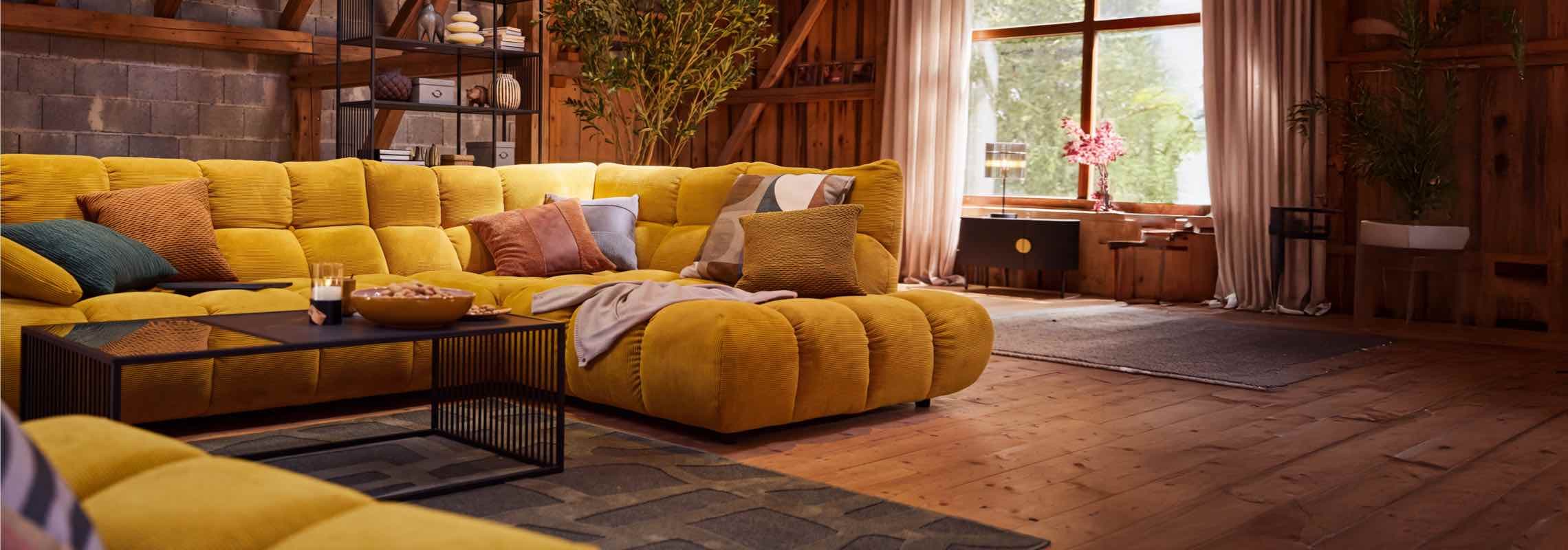 Großes gelbes Ecksofa mit bunten Kissen in rustikalem Wohnzimmer, davor schwarzer Couchtisch mit Snacks und Kerzen
