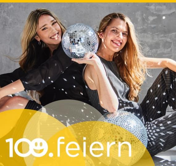 100 feiern – Zwei Frauen sitzen mit Discokugeln am Boden, sie tragen festliche Kleidung und lachen