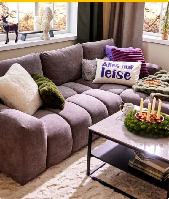 Gemütliches Wohnzimmer mit grauem Sofa, bunten Kissen, Adventskranz und Kerzen auf dem Tisch.