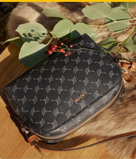 Schwarze JOOP! Tasche mit Muster, umgeben von grünen Blättern und roten Beeren auf einem Holztisch