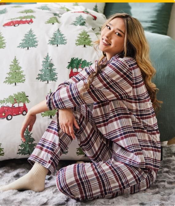 Frau mit langen Haaren sitzt in kariertem Pyjama vor Bett mit weihnachtlicher Bettwäsche mit Tannenbäumen und roten Autos