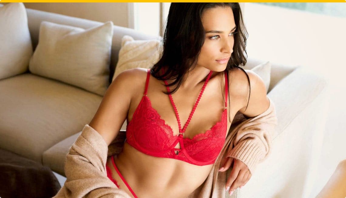 Frau in rotem Spitzen-BH und Slip, trägt beigen Cardigan, sitzt auf Sofa mit Kissen im Hintergrund