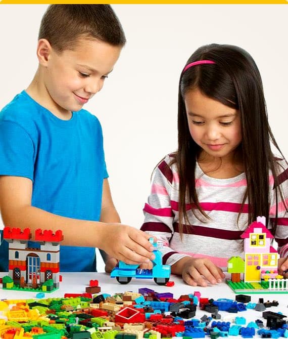 Zwei Kinder spielen mit bunten Bausteinen, bauen Häuser und Fahrzeuge auf einem Tisch.