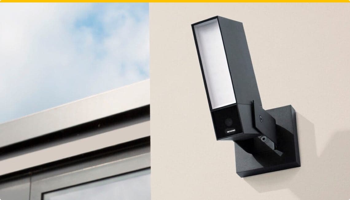 Schwarze Außenleuchte mit Kamera an Hauswand, daneben Teil eines Fensters, blauer Himmel im Hintergrund