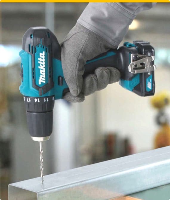 Hand hält Makita-Bohrmaschine, bohrt in Metallstück in heller Werkstattumgebung