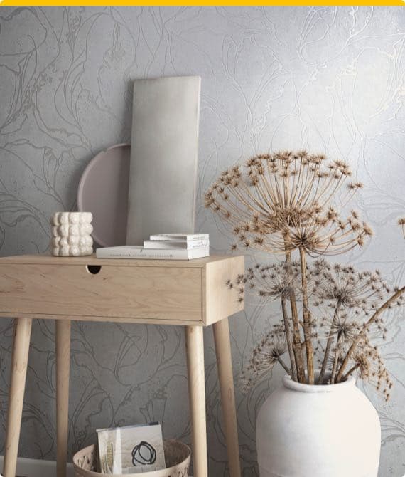 Holztisch mit Dekorationen, darunter eine Vase mit getrockneten Pflanzen, Bücher und ein Kunstobjekt vor grauer Wand